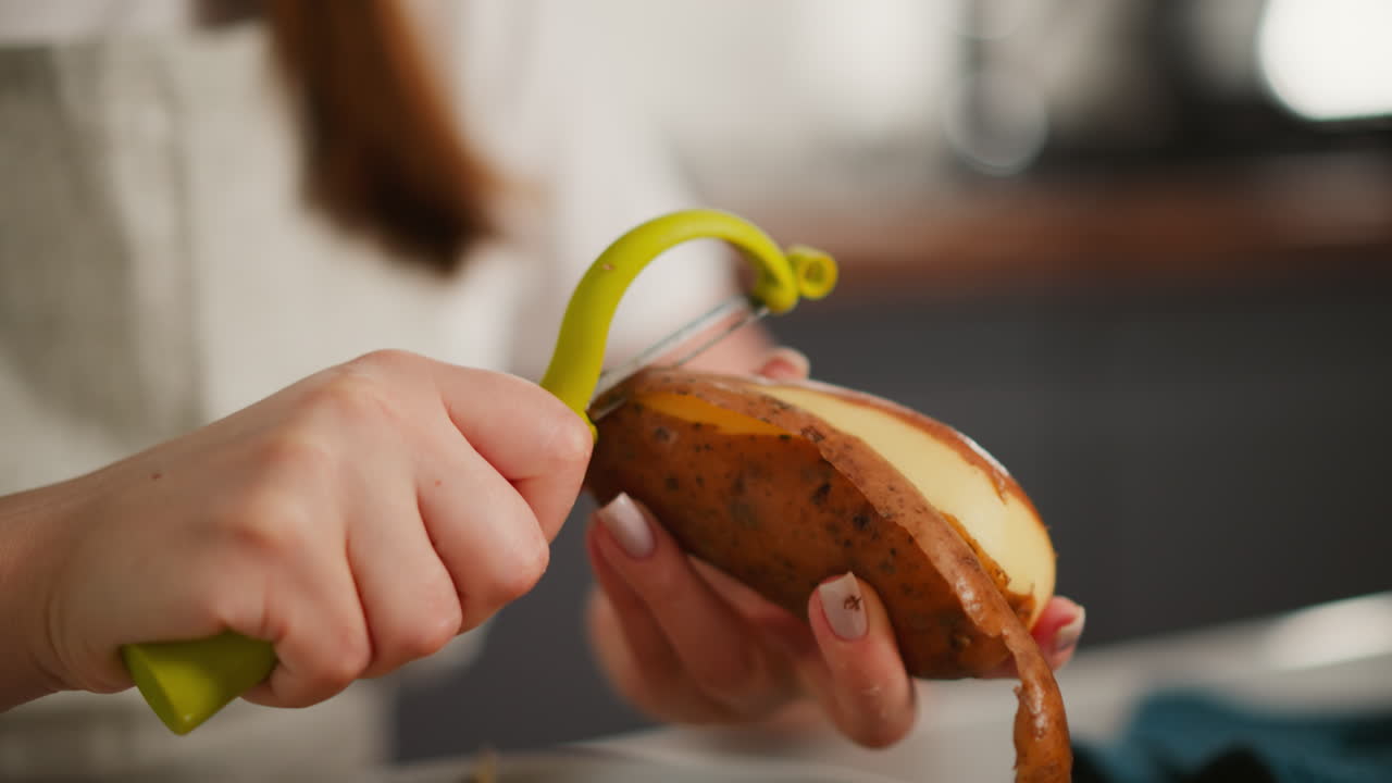 Close up of fair skinned person using yellow peeler to remove potato skin in kitchen setting, hands in focus with blurred background and visible curl of peeled skin hanging from vegetable