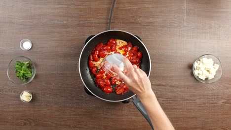 top down shot of pouring milk from glass bowl into large pan with sauteed cherry tomatoes placed on stove on wooden table with other ingredients around