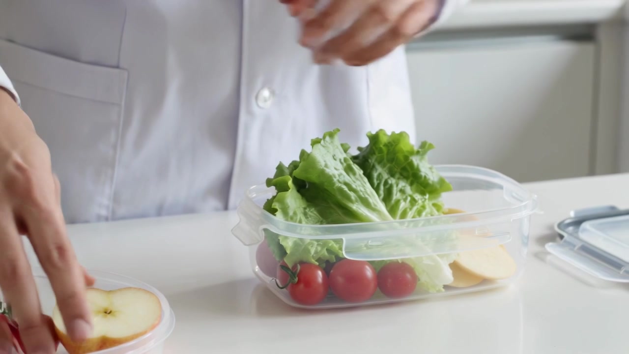 Picking lettuce, person wearing lab coat packing lunch at kitchen counter with clear container