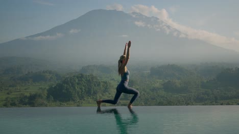 colocar mujer en ropa deportiva de pie en posición de guerrero estirando los brazos por encima de la cabeza, monte agung en el fondo, brillante luz del sol de la mañana