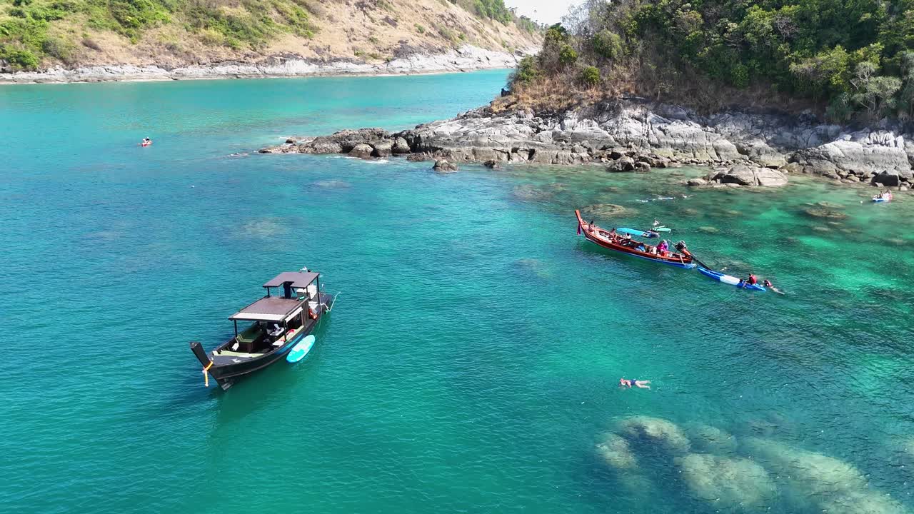 Aerial view of boats and swimmers in clear turquoise waters near rocky shores in Phuket, Thailand, under bright daylight