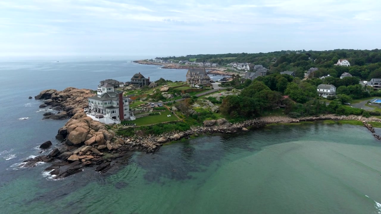Riprese cinematografiche con drone che sorvolano Good Harbor Beach, catturando una spettacolare costa rocciosa, eleganti case sul mare, vegetazione lussureggiante e dolci onde oceaniche sotto un cielo morbido e coperto