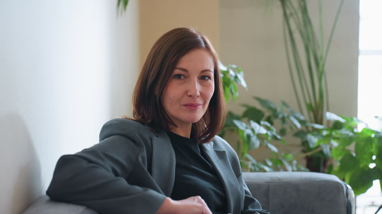 Selbstbewusste Geschäftsfrau im grauen Blazer, die warm lächelnd auf einer Couch in einem modernen Büro mit Zimmerpflanzen und natürlicher Beleuchtung sitzt, was eine ruhige, professionelle Umgebung und ein offenes Auftreten suggeriert.