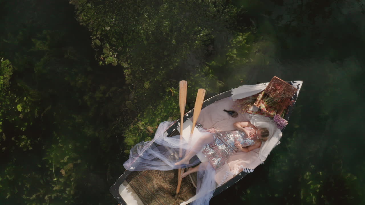 Fashion woman laying in rowing boat putting on sunglasses and drinking champagne on a luxury rowing boat on summer vacation, top down aerial
