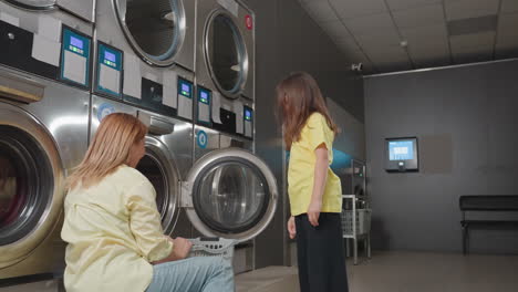 Mother with kid packing clothes into washer at laundromat, woman kneels by open stainless machine, child gestures while basket of towels waits, sorting textiles, clean service routine inside