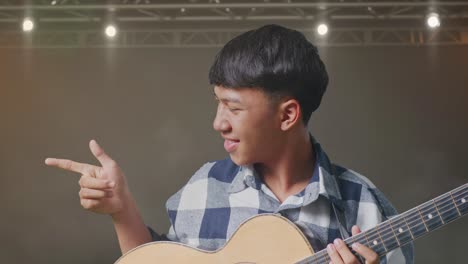 un primer plano de un adolescente asiático con una guitarra sonriendo y señalando hacia el lado en el escenario