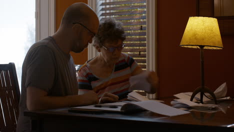 an adult son and his senior mother sit together at a dining table in a warmly lit home, sorting through papers. son helps mother organize receipts, under table lamp light.