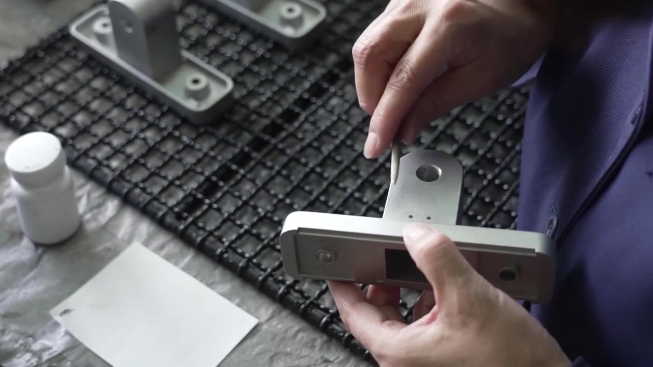 Factory worker inspecting white painted metal finder scope bracket with mounting holes at assembly station