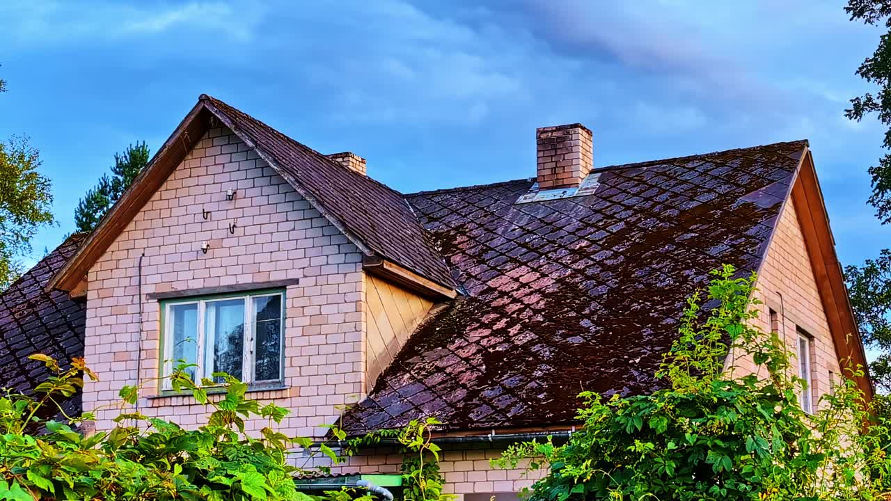 Old brick house with moss-covered tiled roof surrounded by greenery in summer