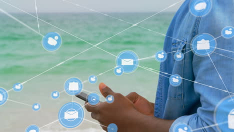 red de iconos digitales contra un hombre afroamericano que usa un teléfono inteligente en la playa