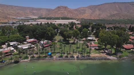 a drone flies back and shows kibbutz ein-gev beach - israel, sea of galilee, the golan heights in the background and people inside the sea of galilee