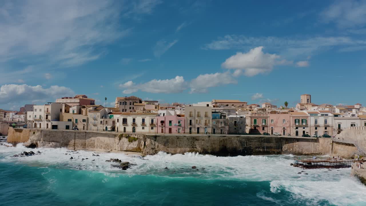 Aerial view of Ortigia island. Flying towards historic old town buildings on a natural fortress by Mediterranean sea with waves crashing on the shore. UNESCO world heritage site in Sicily.