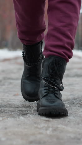 primer plano de las piernas en pantalones de color marrón y botas de diseño negras pisando un camino cubierto de nieve helada, el fondo borroso presenta árboles desnudos y farola