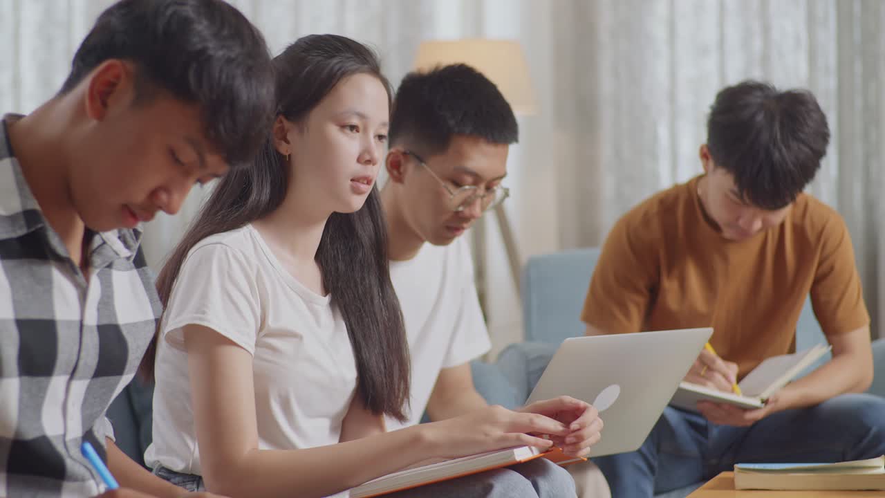 집에서 공부하는 아시안 십대 그룹의 근접 사진. 프로젝트를 하는 데 서로 도움을 주고, 카레드 셔츠를 입은 소년이 노트북에 글을 쓰고, 소녀가 생각하고,  티셔츠를 착용한 소년이 노트북에서 타이핑하고, 다른 소년 에 앉아서 읽고