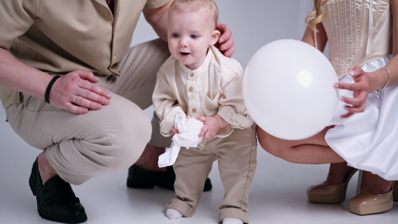 Happy smiling baby holding a toy carries it to camera. Loving parents at backdrop watch their beloved child.