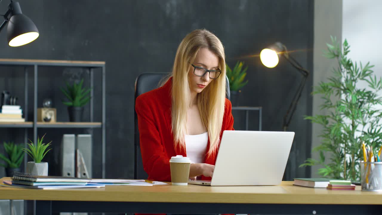 Young Blonde Woman Working Sitting At Desk In Front Of Computer In The Office, Then She Drinks Coffee To Go