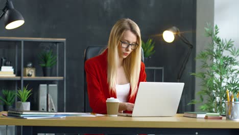 Young-Blonde-Woman-Working-Sitting-At-Desk-In-Front-Of-Computer-In-The-Office,-Then-She-Drinks-Coffee-To-Go