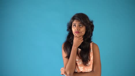 una joven india con vestido naranja que se siente preocupada y mira la cámara y luego piensa en un fondo azul aislado