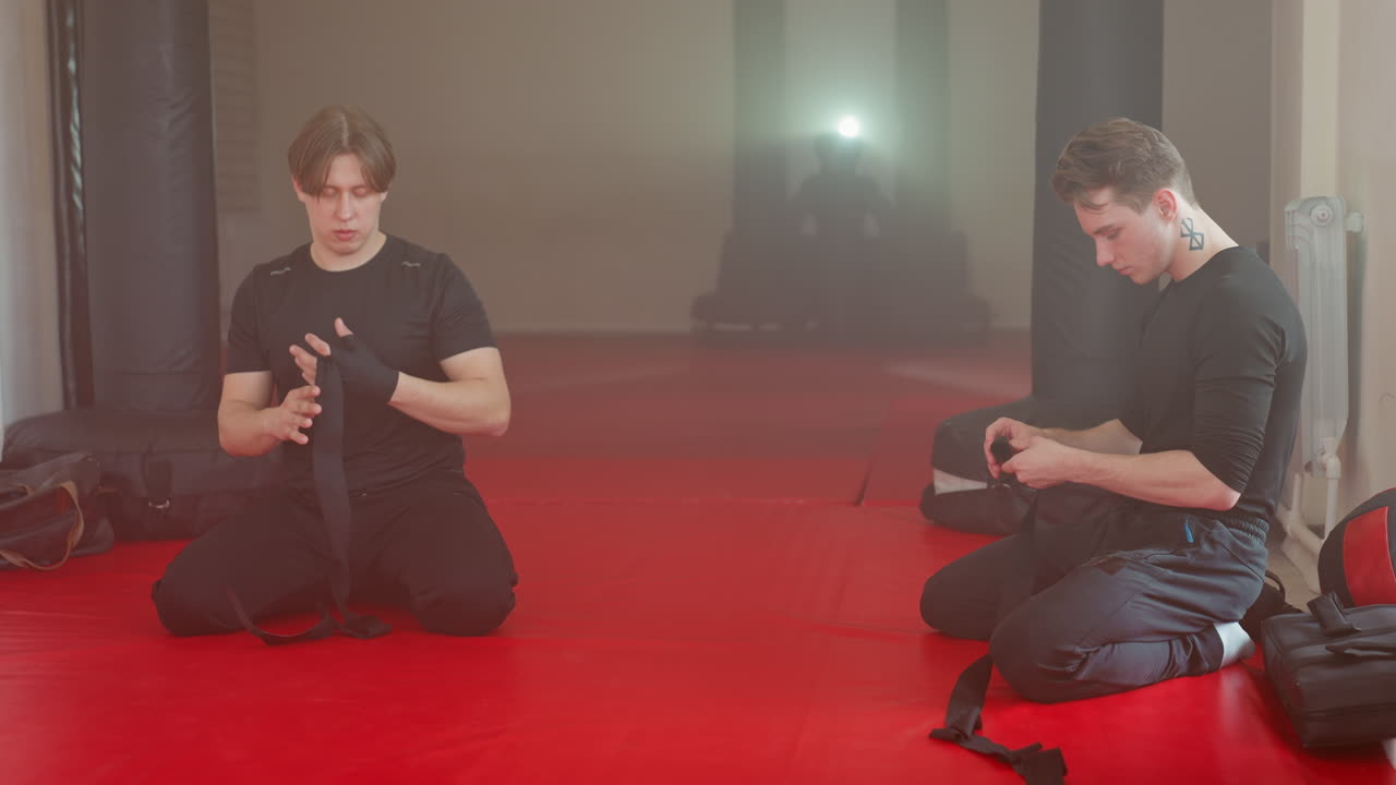 Two athletes kneel on red mat in gym wrapping hands with black straps before training, preparing with focus and discipline, showing strength, determination in martial arts combat sports environment