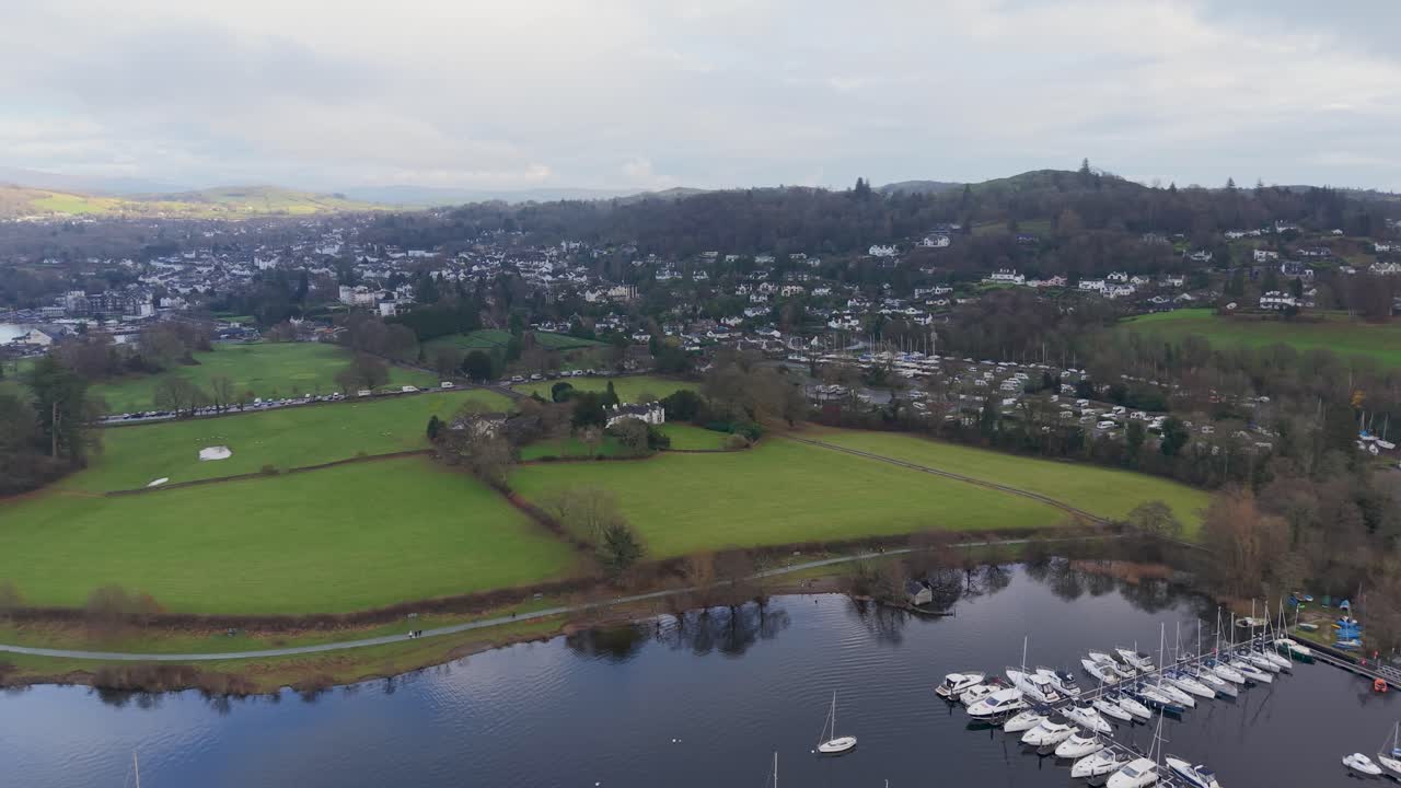 Drone melakukan pan di atas Danau Windermere menuju Bowness, melewati sebuah marina kecil. Pepohonan musim dingin dan kota Bowness berada di latar depan saat puncak bukit bersinar di bawah sinar matahari yang menembus awan