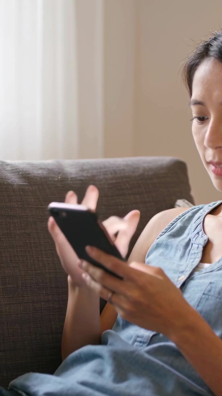 Vertikální video: Žena v modrém topu drží chytrý telefon, ťuká a posouvá na pohovce doma, s prostorem pro text.