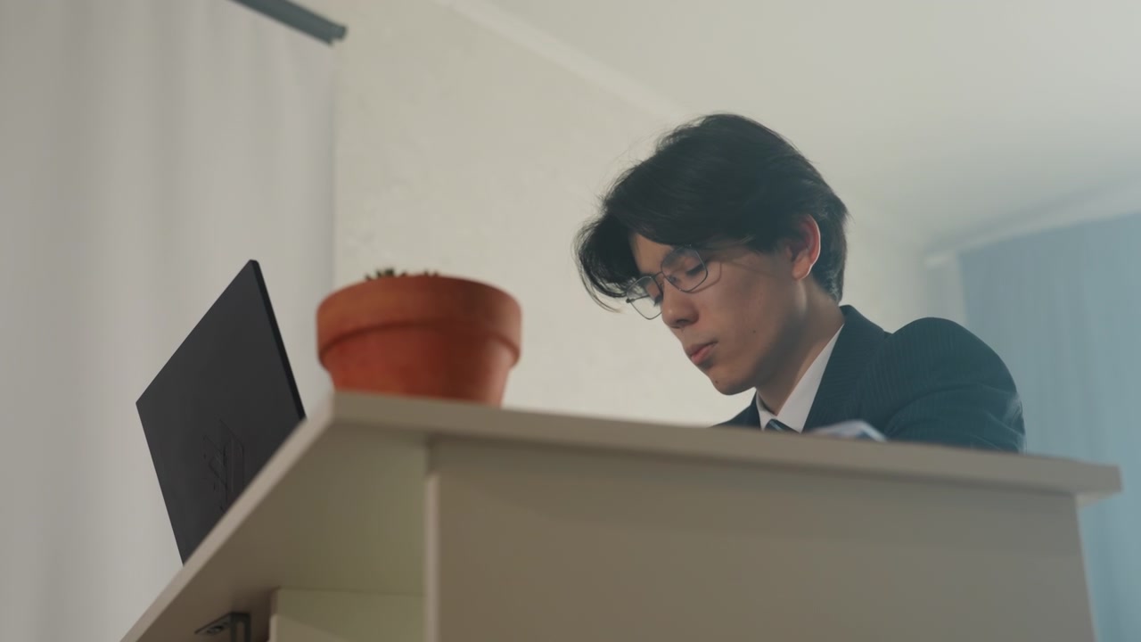 ローアングルからのシネマティックなショットで、ノートパソコンに身を乗り出すアジア人アナリスト。デスクには観葉植物があり、スーツと眼鏡を着用してスプレッドシートとメモを確認している。集中した表情で、暖かく柔らかな照明の中、締め切りが迫る。