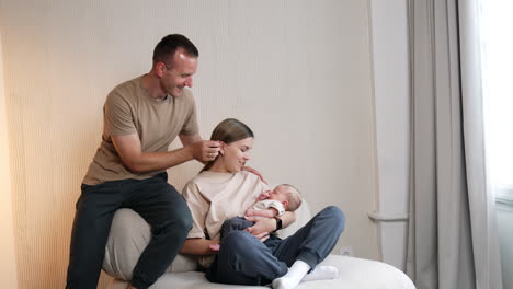 long-haired woman sits on sofa holding her newborn baby. man sitting beside kisses her on the head.