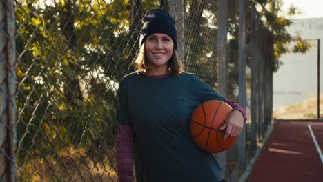 Portrait-of-a-happy-blonde-girl-in-a-black-hat-and-sports-uniform-who-holds-an-orange-basketball-and-leans-on-the-bars-on-a-street-red-court-in-the-morning