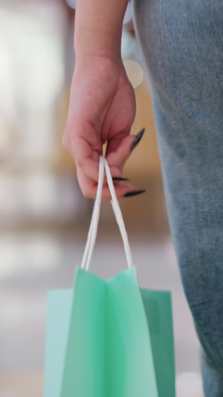 vista de cerca de la pierna de una persona con vaqueros que lleva una bolsa de compras verde mientras camina en el interior, el fondo borroso presenta figuras indistintas, pisos pulidos que reflejan luces suaves