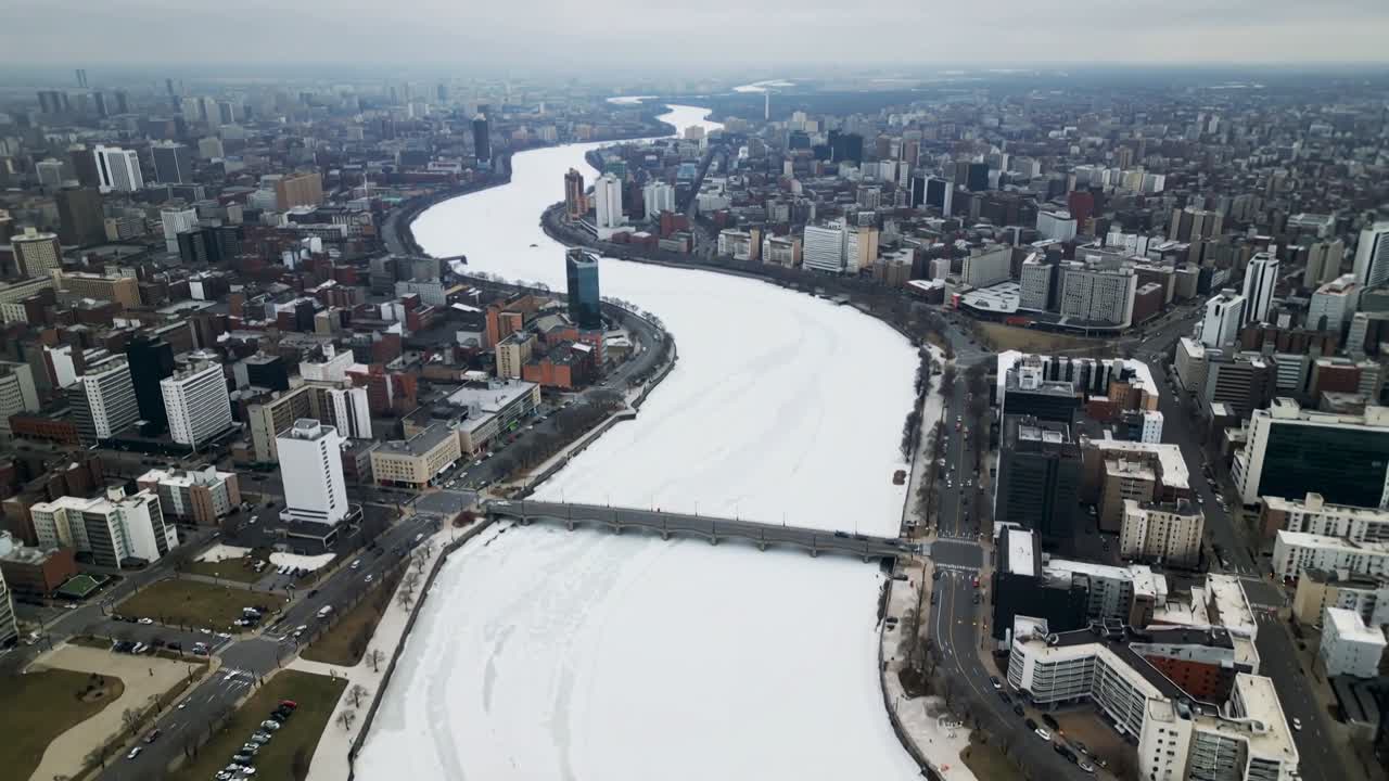 Telecamera drone che avanza lungo un fiume ghiacciato sopra il centro città, mostrando un ponte e una torre di vetro.