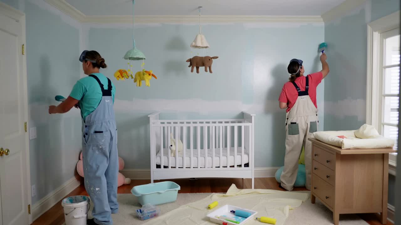 Painting two painters in overalls painting nursery walls, preparing room for baby with crib rollers