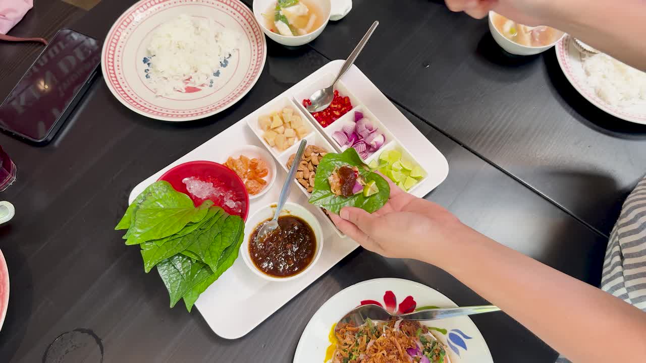 Hands prepare traditional Miang Kham wraps using betel leaves and assorted fillings on a modern dining table, captured in bright, overhead lighting