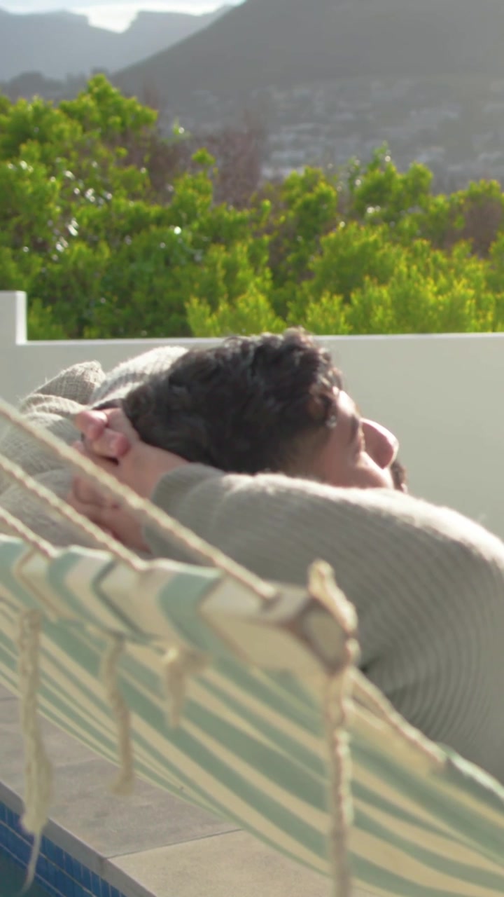 Vertical video: Reclining man in sweater shifting head as sunlight crosses face on terrace, hammock