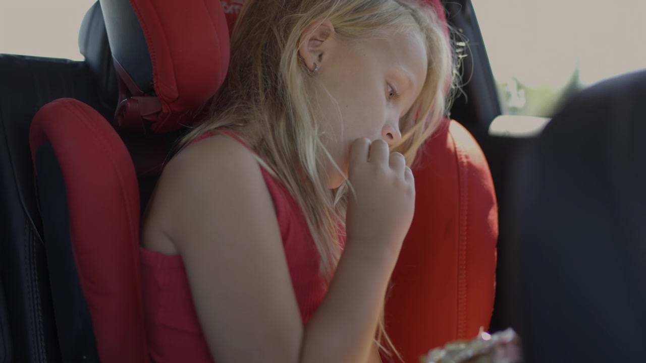 Un niño sentado en una silla de coche, masticando un aperitivo. El niño parece concentrado y contemplativo mientras viste una camiseta roja, rodeado de cojines de asiento rojos