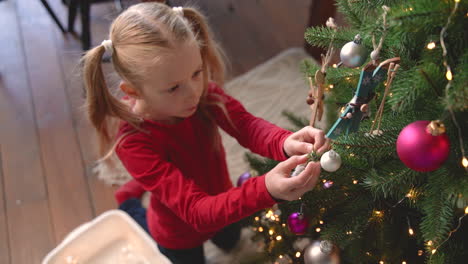 top view of little blonde girl de kneeling on the floor picking up christmas decoration from a box and hanging it on christmas tree 2