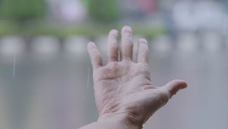 female hand reaching out her hand to feel the rain drops, under heavy rain close up. catches rains on palm, rainy season in slow motion, with urban street background, touch nature, wet hand from rain