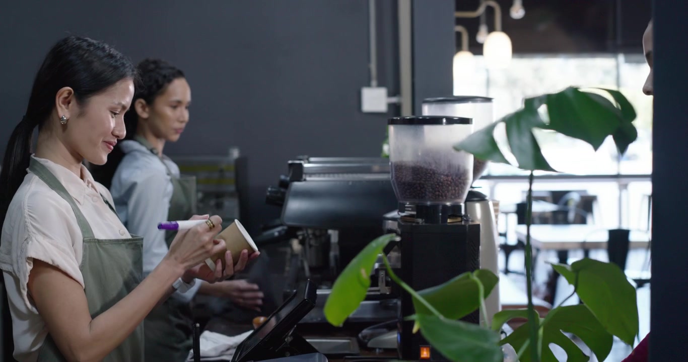 Baristi diversi con grembiuli verdi si voltano mentre una donna alza la mano per ordinare un caffè al bancone, segnando la tazza.