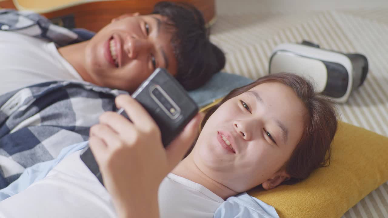집에서 바닥에 카에 누워있는 아시아 십대 커플이 스마트폰으로 비디오 통화를 하고 있습니다. 손을 흔들고, 말하고, 웃고 있습니다.