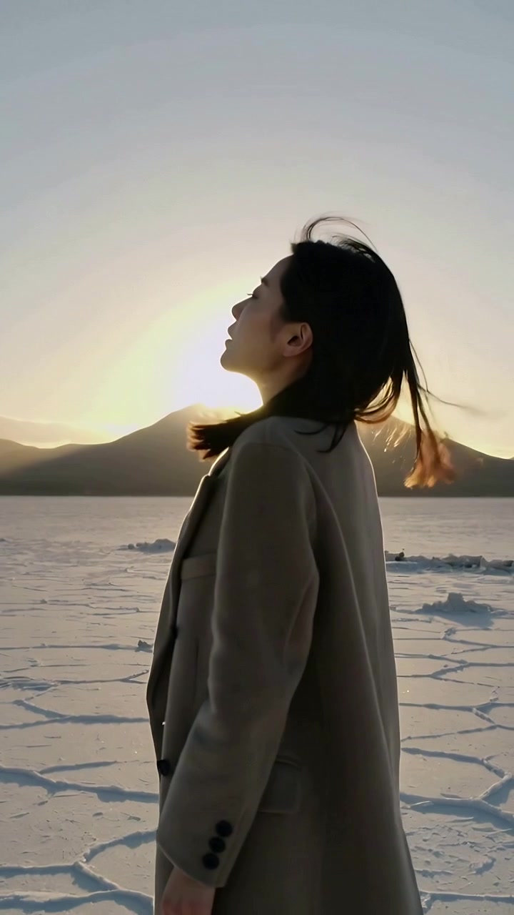 Video verticale: Donna che cammina con cappotto di lana, inclinando la testa verso il sole basso che la disegna su una pianura screpolata.