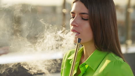 brunette girl in green shirt smoking hookah. beautiful lady breathing out thick white smoke. close up. sunny blurred backdrop.