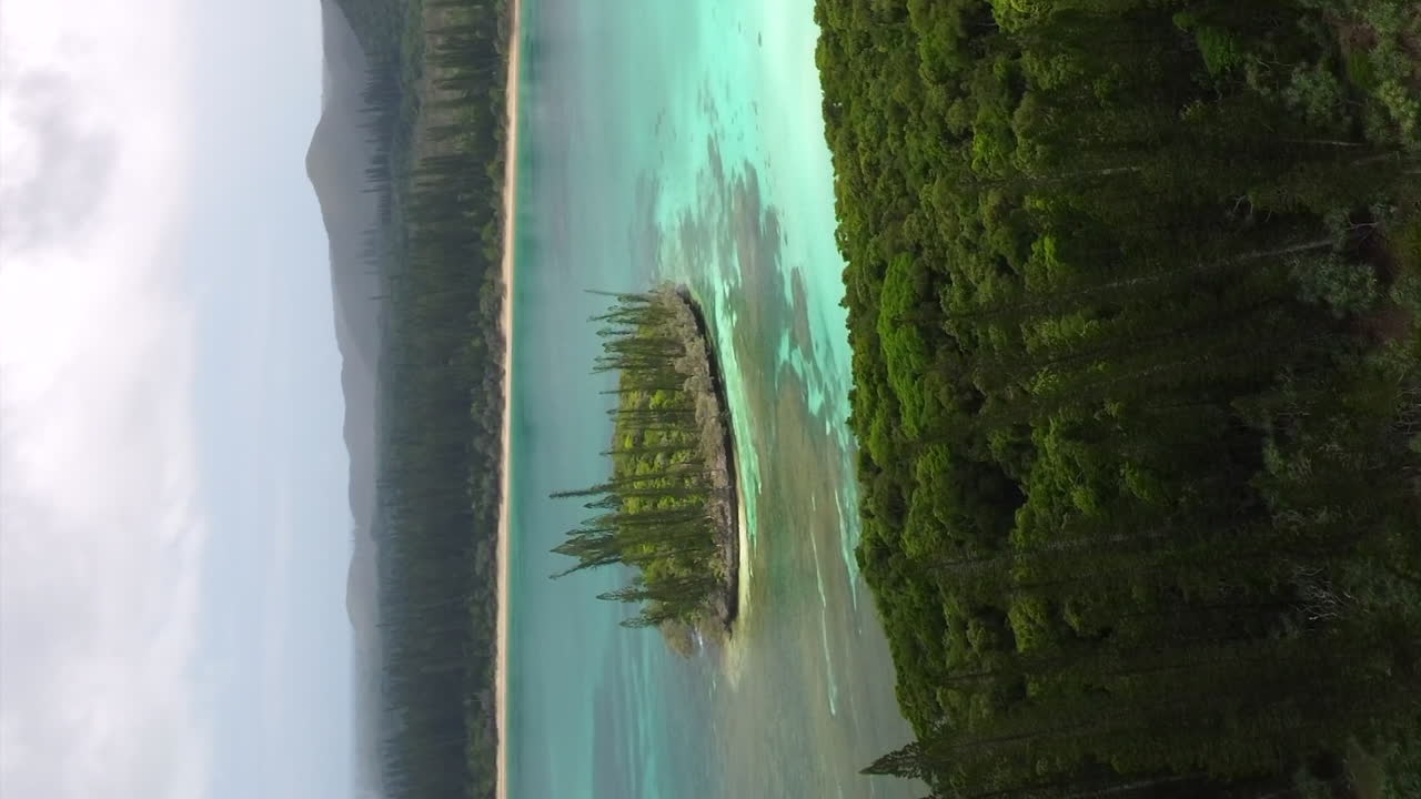 Ripresa aerea ascendente che rivela pini colonnari, acqua cristallina e il picco N'Ga sull'Isola dei Pini.