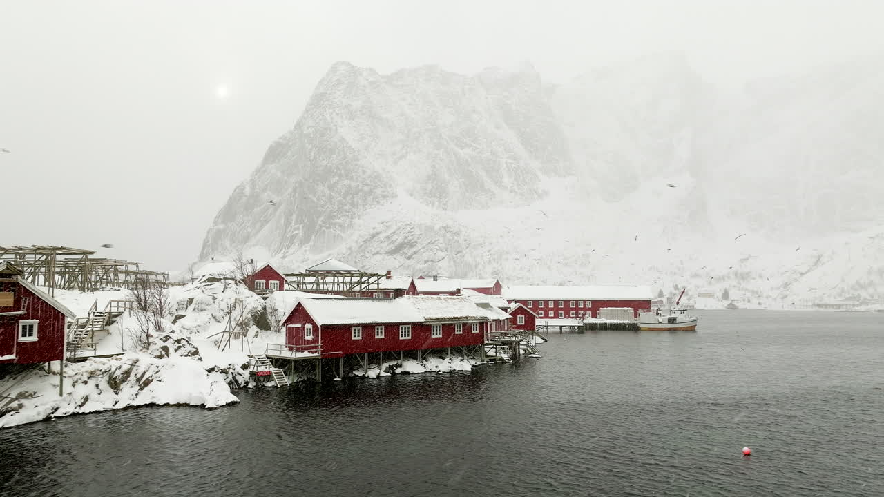 Tradisjonelle rødmalte fiskerhus og tørrfiskstativ langs kystlinjen i Reine, tåkete forhold i det snødekte landskapet.