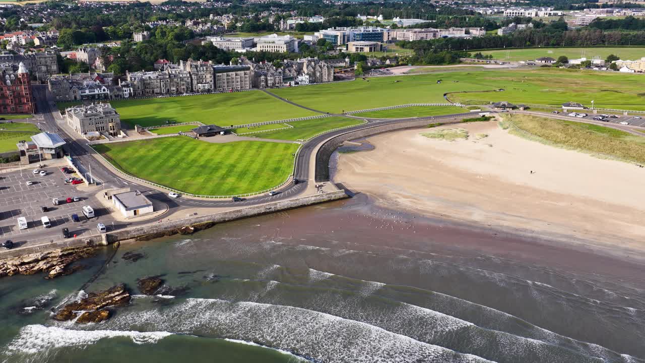 Sweeping aerial shot over golf course, sandy beach, and coastal town under bright daylight