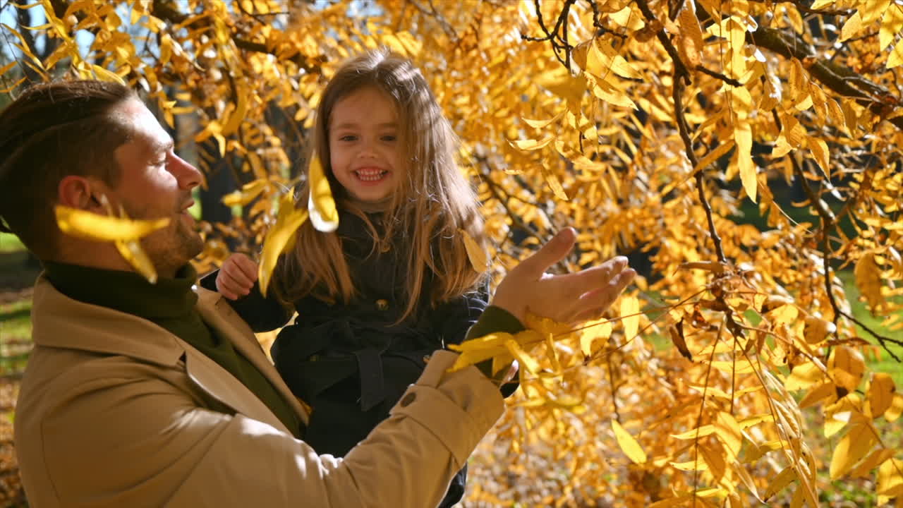 Happy family in an autumn park. Father took his daughter in his arms, collecting leaves from a yellowed tree. Slow motion
