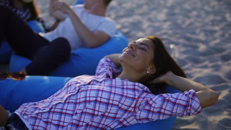 happy young woman laying on easy chair and smiling among group of friends on the beach during a sunset. slowmotion shot