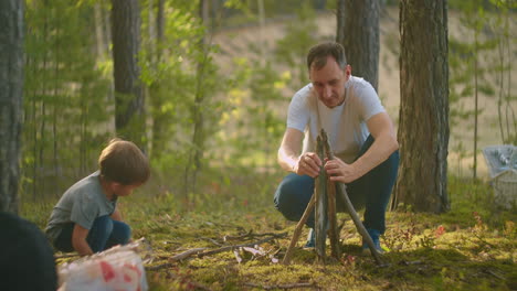 a man in the woods collects and sets up campfire sticks at sunset during a family camping trip