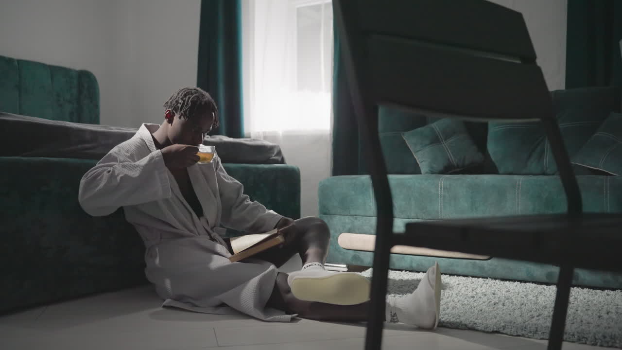 Man in white robe, enjoying cup of tea while sitting on rug in cozy bedroom. Soft morning light pours through window, creating a tranquil and peaceful atmosphere for a relaxing moment