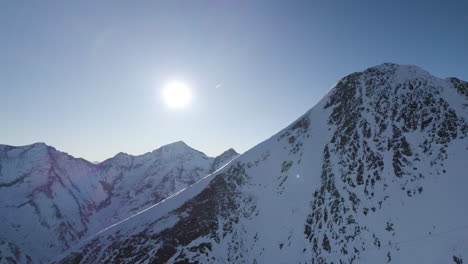 drone panning around austrian mountain peak revealing bright sun with mountain range in background