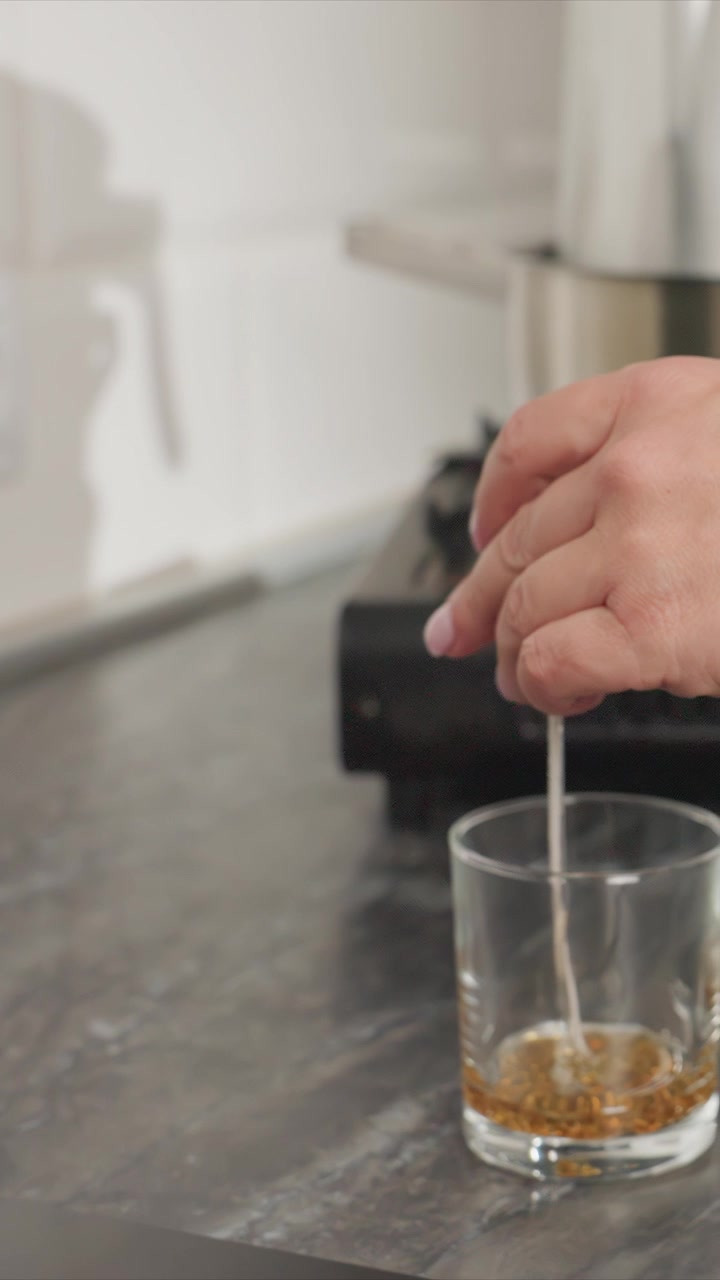 Primo piano di una mano che mescola un liquido in un bicchiere sul bancone della cucina. La scena mostra l'interazione tra mani umane e oggetti, concentrandosi su movimenti precisi in un ambiente interno con luce soffusa.