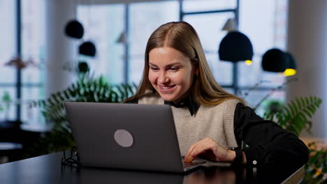 Resilient smiling young woman working on her computer. Happy lady is satisfied with some result and makes a success gesture. Blurred backdrop.